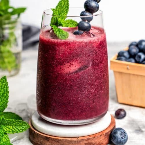 a variety of garnishes including fresh berries, edible flowers, and mint sprigs, arranged around a glass of Violet Drink