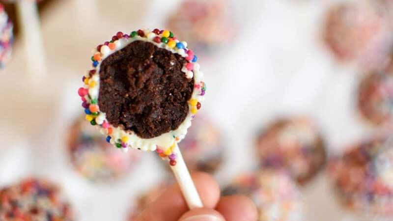 hands dipping cake pops into melted chocolate and decorating with sprinkles