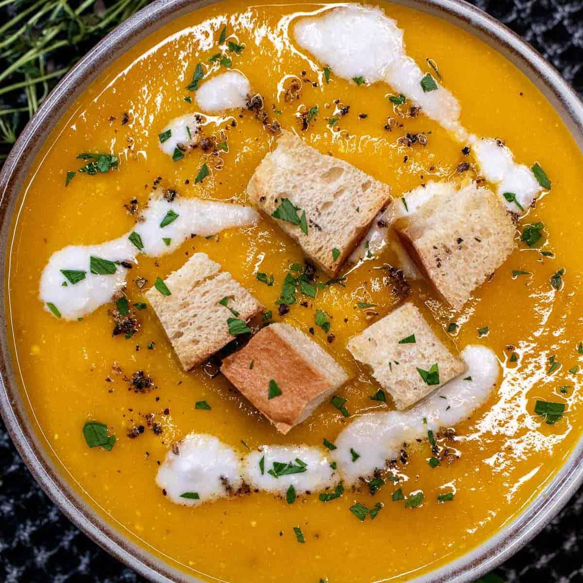 Rustic bowl of creamy roasted carrot and thyme soup garnished with fresh herbs and a swirl of cream, on a wooden table with a cozy winter backdrop.