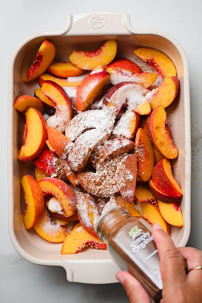 overhead shot of someone sprinkling sugar on sliced peaches in a baking dish