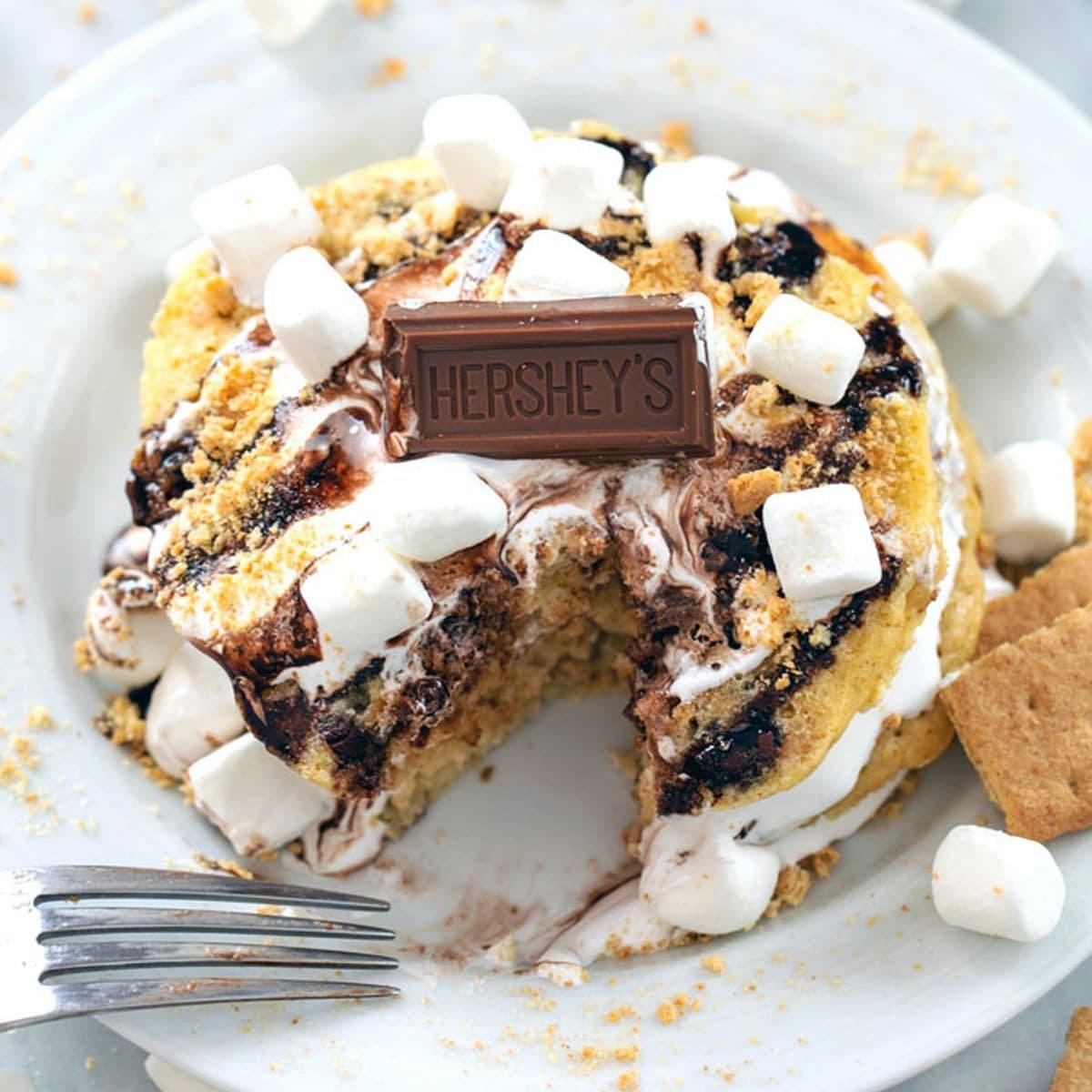 person garnishing s'mores gluten-free pancakes with chocolate sauce and marshmallow fluff