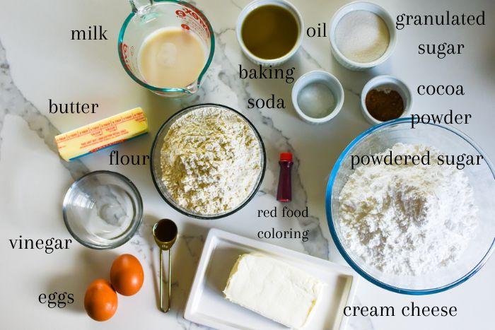 Ingredients for a ruby velvet cake laid out on a wooden countertop, including flour, sugar, cocoa powder, eggs, buttermilk, red food coloring, vanilla extract, and cream cheese