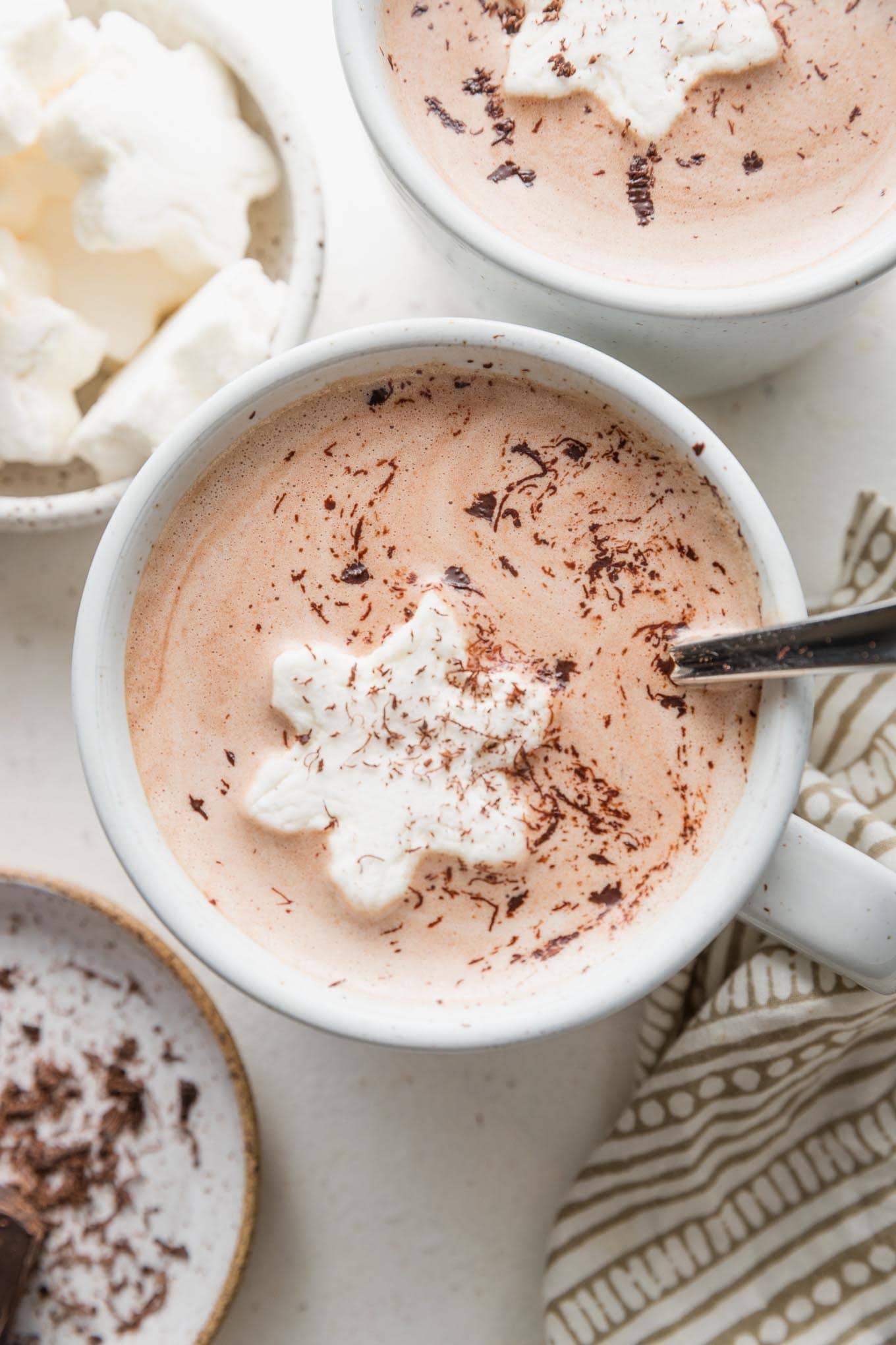 Steaming mug of creamy hot chocolate with generous whipped cream and chocolate shavings on top, cozy setting