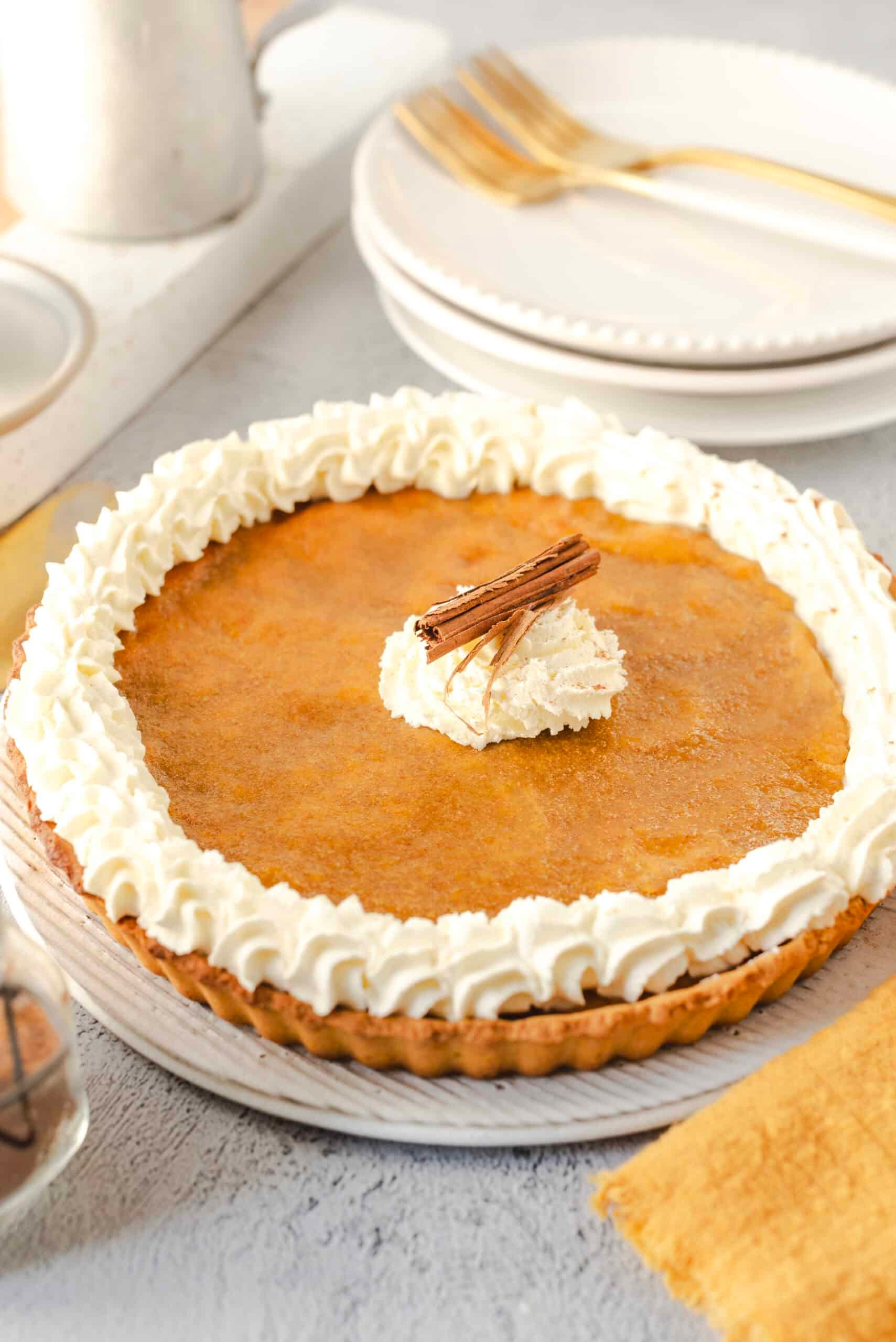 A warm, perfectly baked bakery-style pumpkin pie with a golden, flaky, crimped crust, garnished with whipped cream and a sprinkle of cinnamon, on a rustic wooden table with autumn leaves.