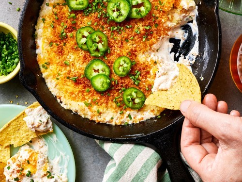 Rustic baked jalapeño popper dip with golden brown topping and fresh jalapeños, surrounded by dippers like tortilla chips and crackers on a wooden table.