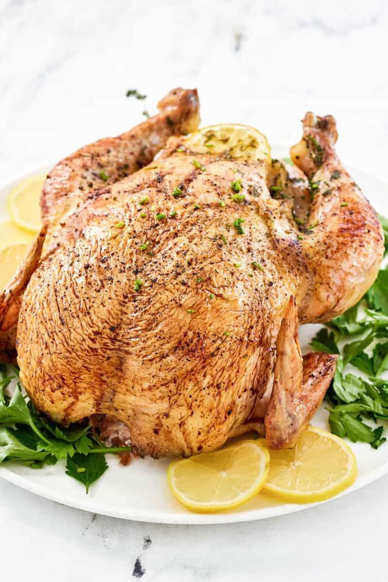 whole golden roasted chicken on a platter with herbs and lemon slices