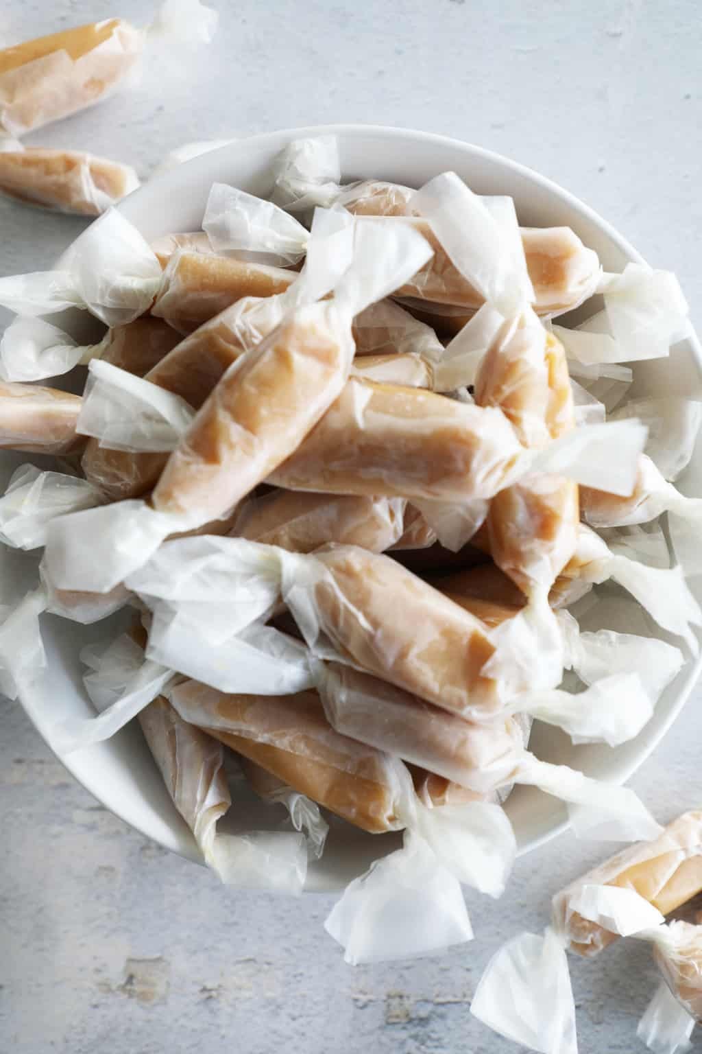 close-up shot of homemade soft vanilla caramels stacked in a rustic bowl, with a few wrapped in parchment paper, natural light