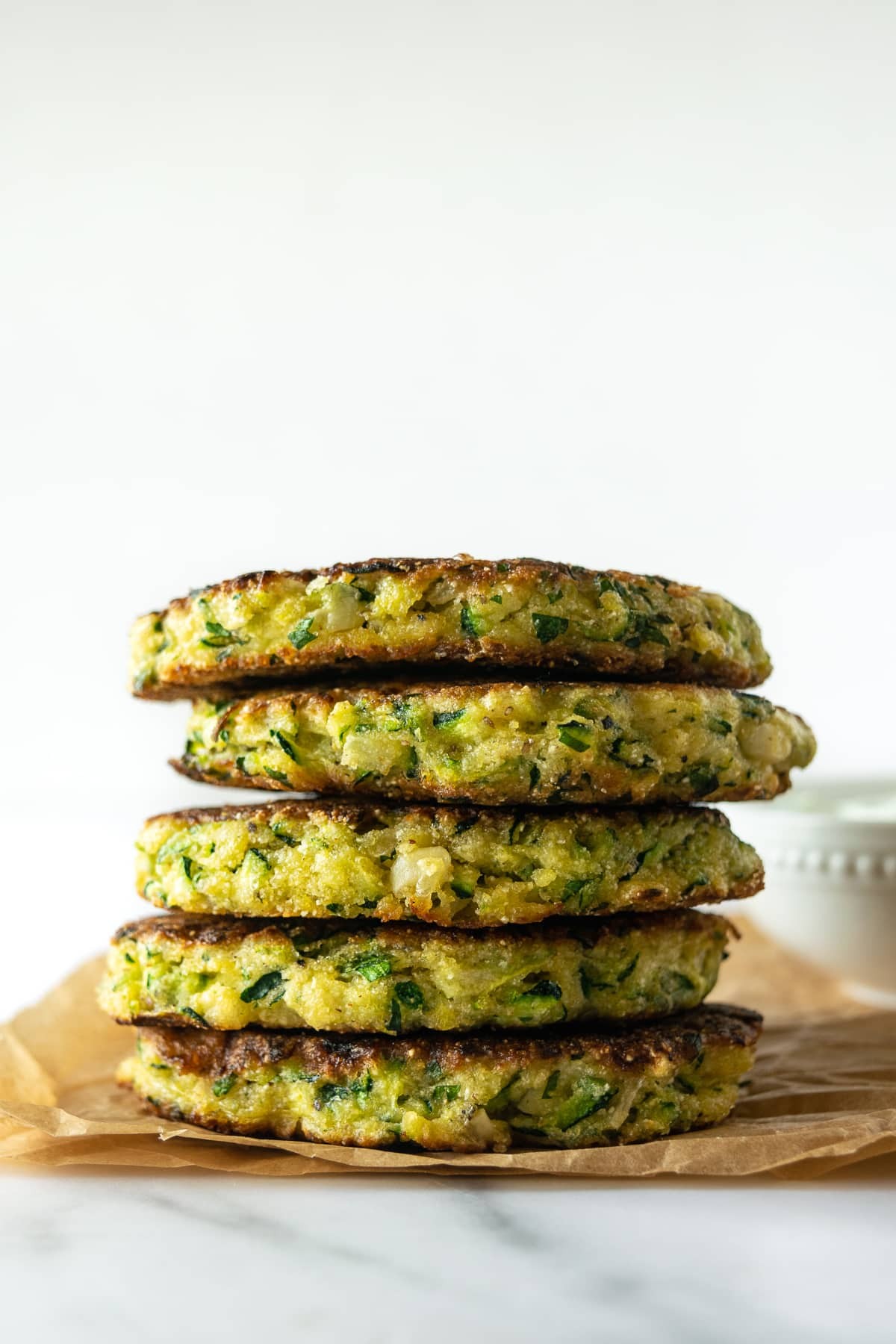 golden baked zucchini fritters stacked on a plate with a side of creamy herb yogurt dip, fresh herbs garnish