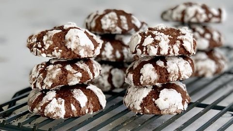 A close-up shot of perfectly baked chocolate crinkle cookies with prominent snowy white cracks on a cooling rack, some stacked, some on a festive plate, warm lighting