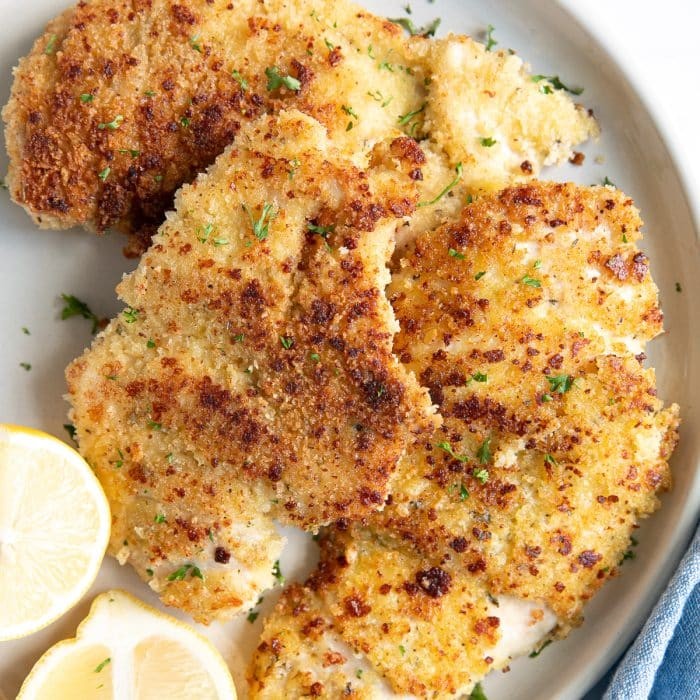 Crispy golden brown parmesan chicken cutlets on a white plate with lemon wedges and fresh parsley, close-up, shallow depth of field, natural light