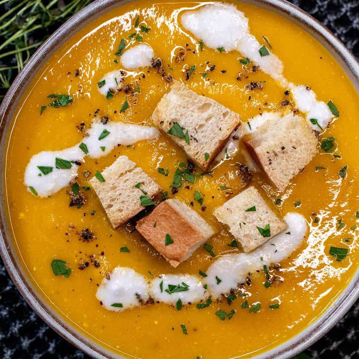 Rustic bowl of creamy roasted carrot and thyme soup garnished with fresh herbs and a swirl of cream, on a wooden table with a cozy winter backdrop.