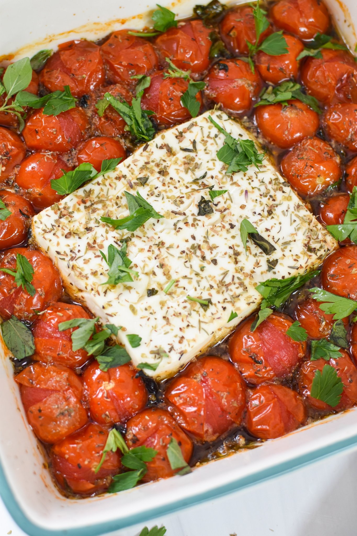 Baked feta cheese with cherry tomatoes and fresh herbs in a baking dish, drizzled with olive oil, close up