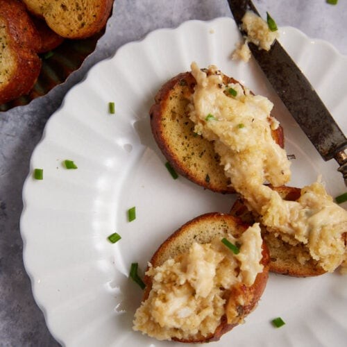 Close-up of golden brown bubbly baked crab dip in a small white ramekin, surrounded by toasted baguette slices and fresh chives.