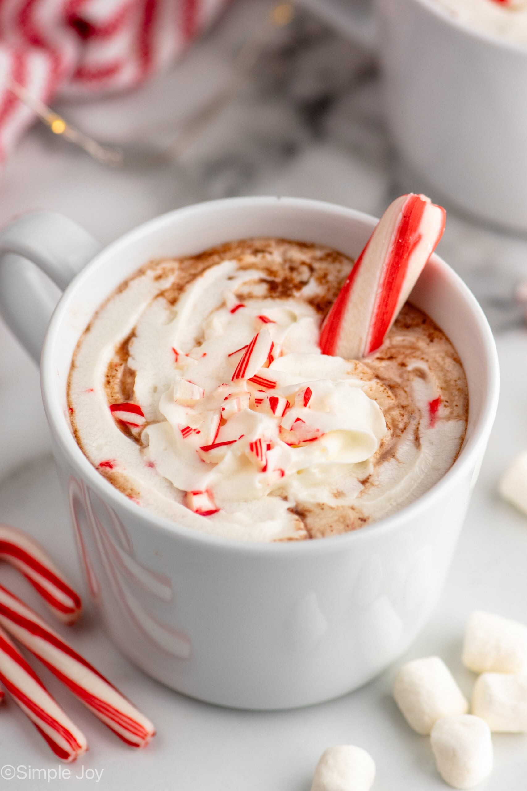 A steaming mug of peppermint hot chocolate garnished with whipped cream and a candy cane, with a subtle frosty or cool background element.